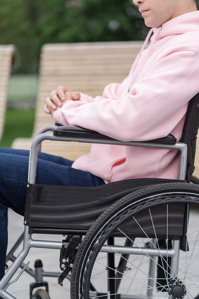 A young adult in a pink hoodie sits in a wheelchair outdoors, showcasing inclusivity and accessibility.